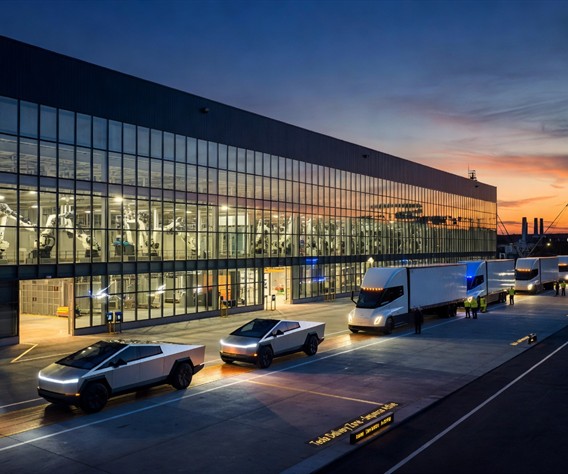 A photorealistic wide-angle view of a gleaming Tesla Gigafactory at dusk, with a line of silver Cybertruck vehicles and white Tesla Semi trucks rolling off the production line onto an illuminated delivery pad, industrial robots visible through massive glass factory walls, warm amber and electric blue lighting, cinematic atmosphere, sense of massive industrial scale and technological precision