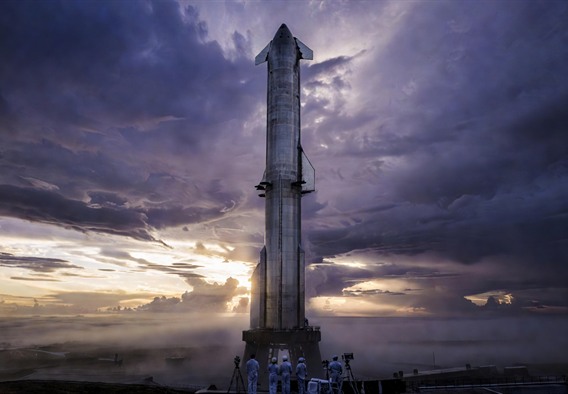 A colossal Starship rocket standing on a launch mount at dawn, surrounded by low coastal fog, with a stormy amber and purple sky in the background. The rocket's stainless steel surface reflects the light dramatically. In the foreground, tiny human silhouettes stand near technical equipment, emphasizing the vehicle's overwhelming scale. Ultra-realistic photography style, cinematic wide angle, deep shadows and high contrast lighting.