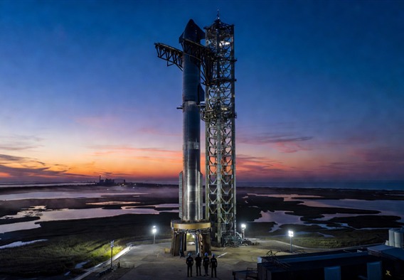 Photorealistic wide-angle dawn scene at a futuristic rocket launch facility on the Texas Gulf Coast, a massive 120-meter stainless steel rocket fully stacked on a towering orbital launch mount, bathed in floodlights against a deep indigo pre-dawn sky streaked with orange on the horizon, small human figures in dark technical clothing visible at the base for scale, salt marshes and shallow water reflecting the lights, cinematic atmosphere, hyper-detailed engineering infrastructure visible