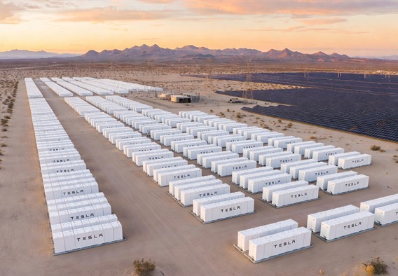 Photorealistic wide-angle aerial view at golden hour dawn of hundreds of large white Tesla Megapack battery storage units arranged in precise geometric rows across a flat desert plain, flanked by vast utility-scale solar panel arrays catching the first rays of sunrise, distant mountains silhouetted against an orange-pink sky, power transmission lines converging at a substation in the background, conveying immense scale and the quiet power of clean energy infrastructure