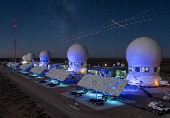 A sweeping nighttime panoramic view of a next-generation satellite ground station facility, multiple large white radome structures and flat phased-array panels arranged across an open landscape, glowing blue and white from internal electronics, a dense star field visible above with faint orbital satellite trails streaking across the sky, ultra-realistic photographic style with dramatic lighting, cinematic composition emphasizing the scale and futuristic nature of the infrastructure